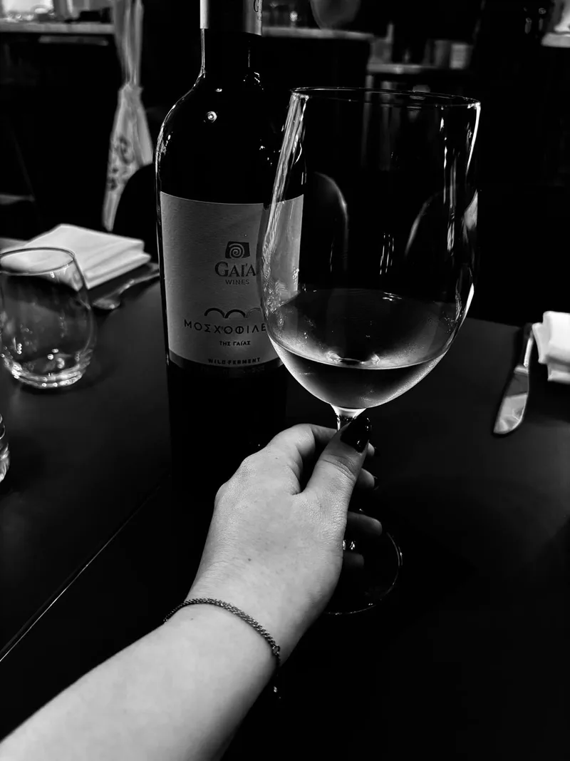 A black and white photograph of a hand holding a wine glass beside a bottle