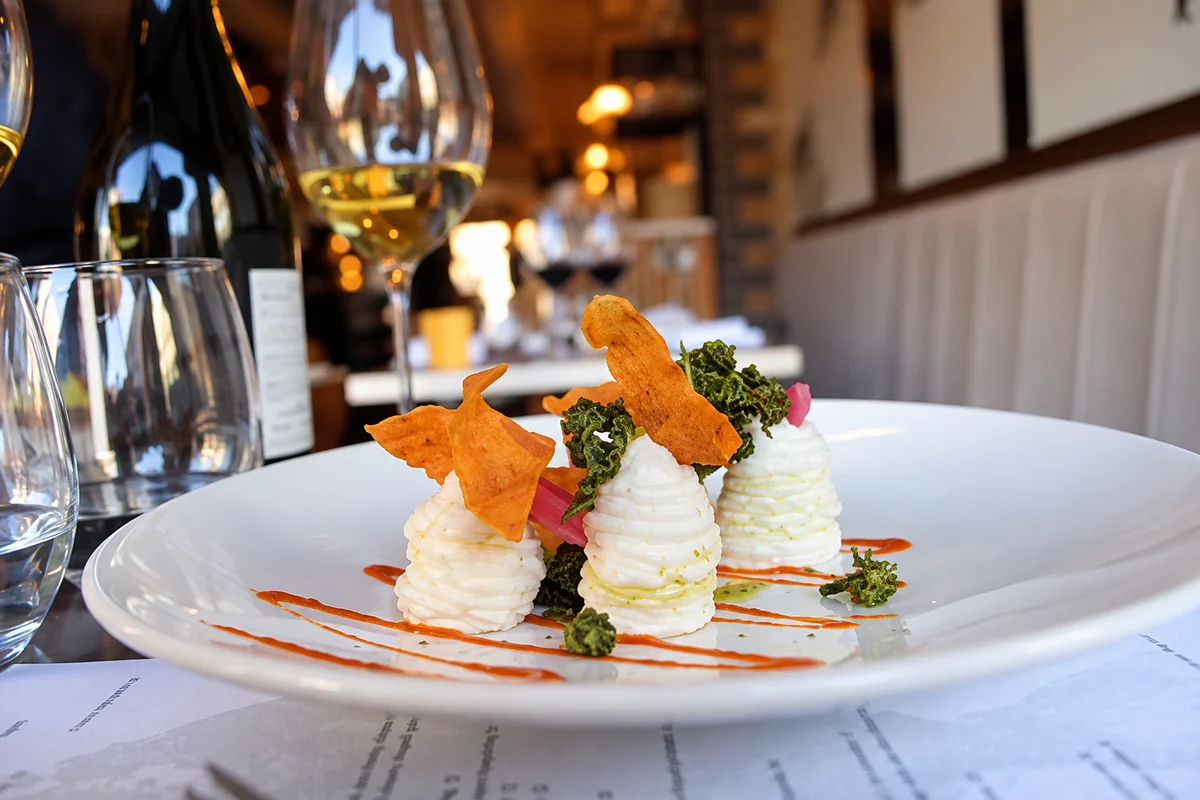 A plated dish with orange and green garnishes, with wine glasses and the restaurant interior behind